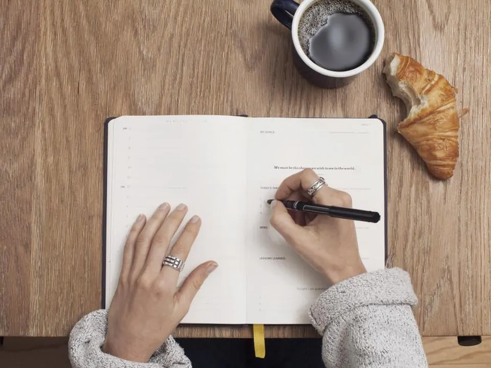 Hands writing in a notebook next to coffee and a croissant